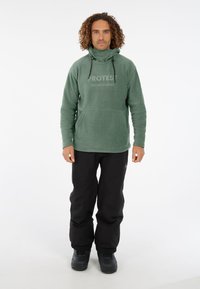 Groene fleece hoodie met hoge kraag, voorzien van een buidelzak en een logo-opdruk. Gecombineerd met zwarte geïsoleerde broek en laarzen.
