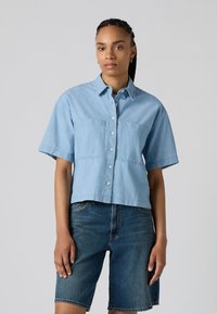 Camicia azzurra a maniche corte con bottoni, dotata di due tasche sul petto, design cropped e vestibilità comoda, abbinata a pantaloncini di denim.