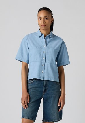 Light blue short-sleeve button-up shirt with two chest pockets, cropped design, and a relaxed fit, paired with denim shorts.
