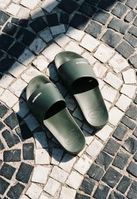 Sandales slides vertes Havaianas posées sur un pavé en pierre à motif noir et blanc avec des contrastes de lumière et d'ombre.