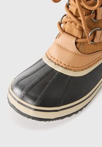 Svart gummistøvel med en strukturert tåhette, overdel i brun skinn og et mønstrete design. Har grå snørebånd og en off-white såle.