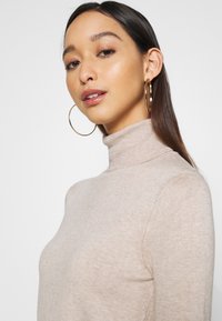 Beige turtleneck-neule pehmeästä ribbatuista langoista, joissa on tiukat hihat. Mallilla on suuret renkaita korvissa; sileät hiukset, joissa on kevyt aalto.