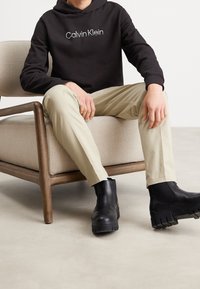 Haut à capuche noir avec le logo "Calvin Klein", pantalon beige clair et bottines noires avec une semelle épaisse et texturée, assis sur un fauteuil de couleur claire.