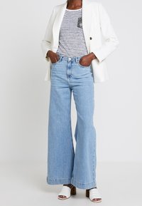 Blazer blanco sobre una camiseta de rayas, combinado con jeans de pierna ancha azul claro y sandalias de tacón blanco. Notables acentos de botones en el blazer.