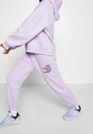 Osoba ubrana w jasnofioletową bluzę z kapturem i dopasowane spodnie dresowe z napisem "The Future Surf Society", idąca w półkroku, na jednolitym tle.