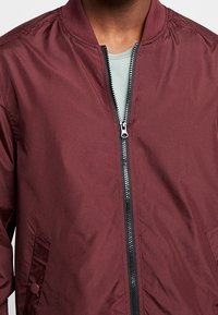 Bordowa kurtka bomber z zamkiem z przodu, żeberkowym kołnierzem i dwoma bocznymi kieszeniami. Wykonana z gładkiego, lekkiego materiału o matowym wykończeniu.