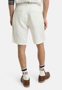 Weiße Leinen-Shorts mit lockerer Passform, ausgestattet mit zwei Gesäßtaschen und einem dezenten Texturmuster. Kombiniert mit braunen Schuhen und gestreiften Socken.