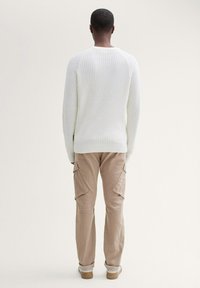 Witte geribde trui met lange mouwen, gecombineerd met beige cargobroek met zijzakken en een ontspannen pasvorm. Achtergrond is neutraal.