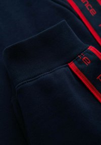 Morska niebieska bluza z długim rękawem i ściągaczem, z czerwonymi przeszyciami oraz częściowym czerwonym tekstem wzdłuż szwu.