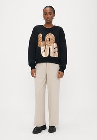 Mulher em pé vestindo sweatshirt preta com texto "LOVE" em lantejoulas douradas, calças largas bege e sapatos pretos robustos sobre fundo branco.