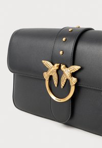 Μαύρη δερμάτινη τσάντα χειρός με χρυσό κούμπωμα σε σχήμα πουλιού, διακοσμητικές λεπτομέρειες με τρουκς στη φλάντζα και λεία υφή. Το λογότυπο της μάρκας είναι ορατό στο κούμπωμα.