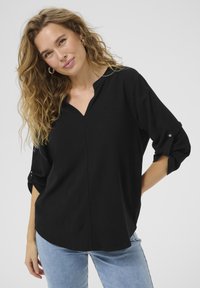 Camicetta nera, morbida e svasata, con scollatura a V, maniche a tre quarti arrotolate e una texture liscia. Indossata con jeans azzurri chiari per un contrasto.