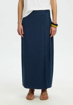 Mujer con una falda larga estilo envolvente color azul marino, top blanco, pulseras amarillas y negras, y sandalias marrones, posando frente a un fondo liso.