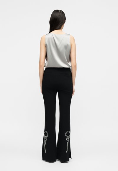 Γυναίκα με μακριά σκούρα μαλλιά που φοράει αμάνικο ασημί τοπ και μαύρο κλασικό παντελόνι με λευκή κυκλική λουλουδάτη κεντητική στο πίσω μέρος των κάτω ποδιών.