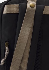Zaino in tessuto nero con una tracolla in oro intrecciato e una cerniera con un tirante decorativo. Pannello in rete scuro visibile.
