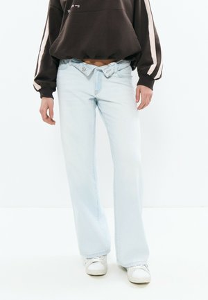 Personne portant un jean large bleu clair avec une taille à double bouton, un sweat-shirt foncé à rayures blanches, et des baskets blanches.