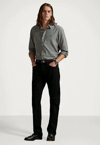 Szaro-biała koszula w kratę zapinana na guziki, czarne spodnie i czarne buty; model stoi z rękami w kieszeniach, nosząc zegarek na nadgarstku.