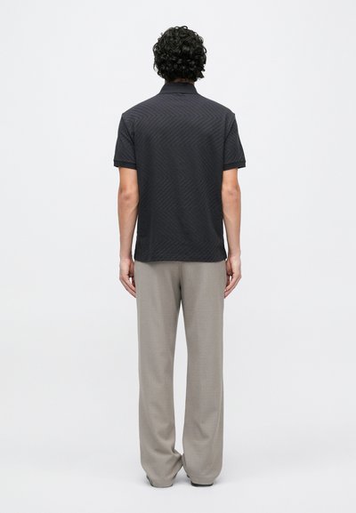 Melna īsroku polo krekls ar teksturētu zivtiņas rakstu, valkāts kopā ar gaiši pelēkiem platām biksēm, skatīts no aizmugures.