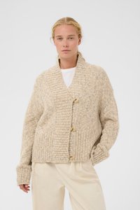 Frau mit blondem Haar, die eine beige, übergroße Strickjacke mit großen Knöpfen und cremefarbene Hose trägt, vor schlichtem Hintergrund.