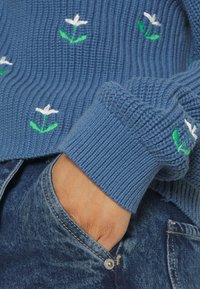 Blå stickad tröja med vit blomstickning och gröna blad. Ribbad struktur på ärmslut och nederkant. Hand vilande i fickan på denimjeans.