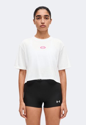 Kvinde med mørkt hår sat tilbage har en hvid cropped T-shirt med pink logo og sorte tætsiddende shorts med hvidt logo, står mod en ensfarvet baggrund.