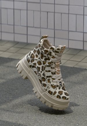 Sneaker med chunky sål i beige og brun leopardprint, hvide snørebånd og mærket tunge, placeret på asfalt nær flisevæg baggrund.