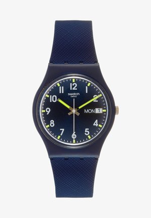 Dunkelblaue Swatch-Armbanduhr mit neongelben Zeigern, weißen Stundenmarkierungen und einer Tagesdatumsanzeige, die Montag, den 1. zeigt.