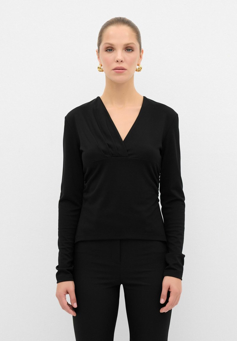 Top nero a maniche lunghe con scollo a V e dettagli di piega frontale, realizzato in un tessuto liscio e con una vestibilità slim. Indossato con pantaloni neri.
