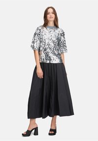 Silberner Pailletten-Oberteil mit kurzen Ärmeln, kombiniert mit einem langen schwarzen Rock. Klobige schwarze Absatzschuhe runden den Look ab. Glänzende Textur durchgehend.