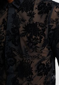 Camicia nera trasparente con motivo floreale e ricami a texture, colletto con bottoni e dettagli sul davanti.