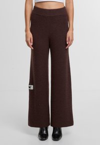 Široké kalhoty z tmavě hnědé texturované tkaniny. Mají vysoký elastický pas a čtvercovou nášivku s logem na levé straně.