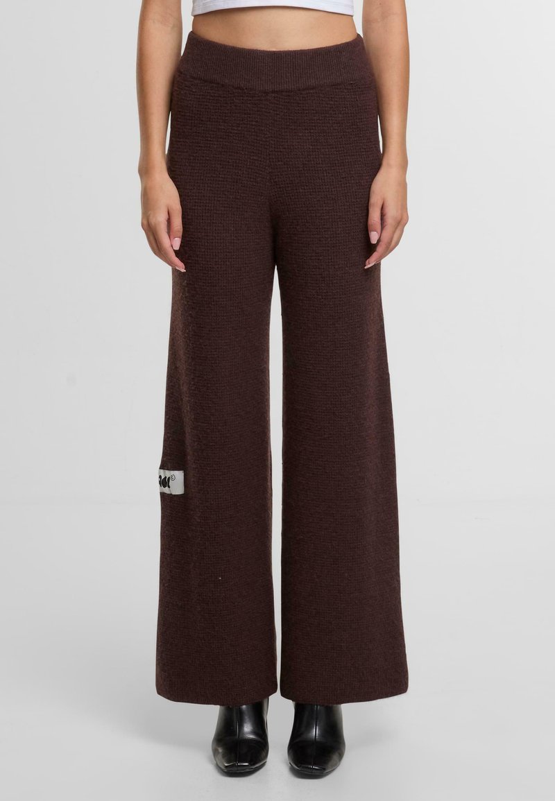 Široké kalhoty z tmavě hnědé texturované tkaniny. Mají vysoký elastický pas a čtvercovou nášivku s logem na levé straně.
