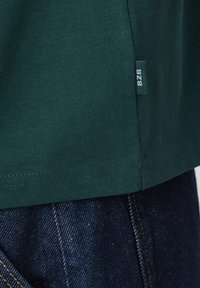 T-shirt en coton vert avec un col ribbed et des coutures visibles à l'ourlet. Présente une petite étiquette tissée avec "B2B" sur le côté.