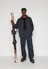 Mann in schwarzer Skijacke, Hose, Stiefeln und Skibrille, der ein Paar Skier aufrecht gegen einen schlichten Hintergrund hält.