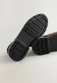 Braune Schuhe mit einer strukturierten schwarzen Gummisohle, die tiefe Rillen für besseren Halt aufweist. Die Seitenansicht zeigt die Nähte des Obermaterials.
