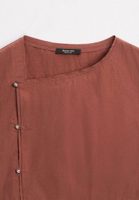 Rostfärgad blus med rund halsringning, som har en ficka och silverknappar längs sidan. Tillverkad av strukturerat tyg.
