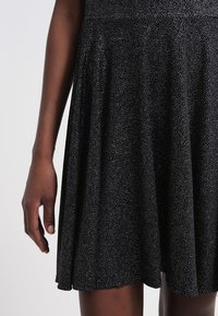 Gros plan d'une personne portant une jupe noire à petits pois blancs, montrant le bas du torse et un bras détendu le long du corps.