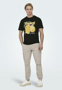Camiseta negra con gráfico de fruta y texto "Florida Tropical". Combinada con pantalones cargo beige y zapatillas blancas. Atuendo simple y casual.
