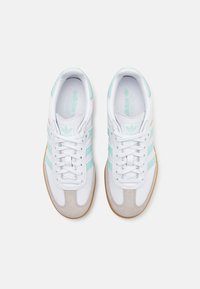 Weiße Sneaker mit hellblauen Streifen, grauen Wildlederakzenten und einer strukturierten Gummisohle. Die Schnürsenkel sind weiß mit einer glatten Oberfläche.