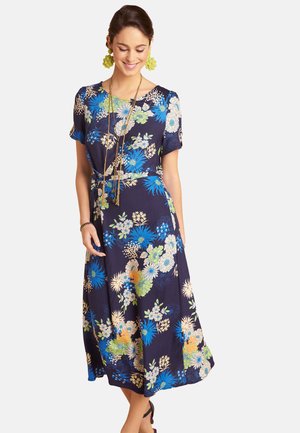 Frau trägt ein marineblaues, knielanges Blumenkleid mit kurzen Ärmeln, grüne Ohrringe, geschichtete Halsketten und ein Armband, lächelt mit geschlossenen Augen.