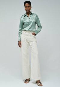 Camisa de satén verde claro de botones con mangas largas, combinada con pantalones de mezclilla blanca de pierna ancha y tacones descubiertos.