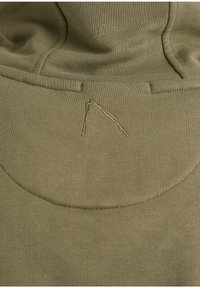 Gros plan d’un tissu vert olive avec une couture courbée et un détail de broderie en forme de V inversé cousu au centre.