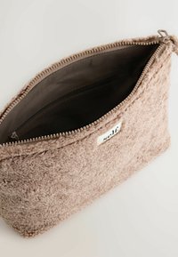 Blød beige fleece kosmetikpung med lynlås åben, som viser mørkt stofforing og lomme med lynlås indeni, mærke med teksten "selv" på ydersiden.