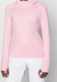 Woman wearing a fitted long-sleeve light pink top with thumb holes and white pants, shown from neck to hips.