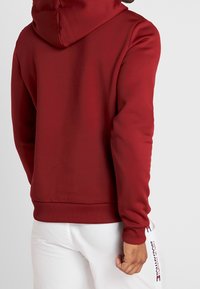 Rode hoodie met een zachte textuur, voorzien van een grote capuchon, strakke manchetten en een tailleband. De achterkant toont een effen kleur met minimale details.