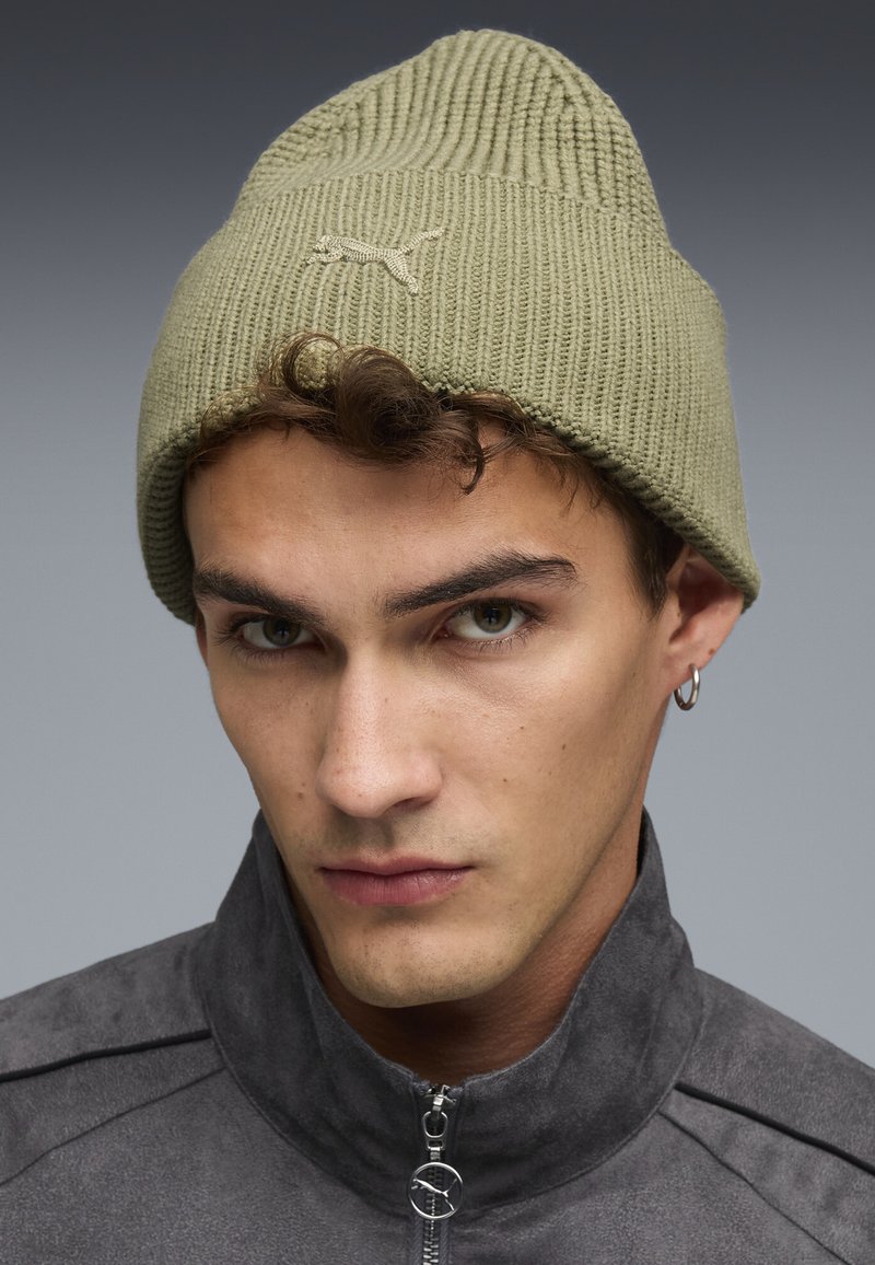 Cappello di maglia verde oliva con una texture a coste e un piccolo logo ricamato. Indossato con una giacca grigia con zip caratterizzata da una finitura liscia.