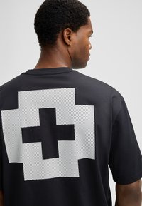 Mann, der sich abgewandt hat, trägt ein schwarzes T-Shirt mit einem großen, weißen, pixelierten Kreuzdesign auf dem Rücken. Ein goldener Creolenohrring ist am linken Ohr zu sehen.