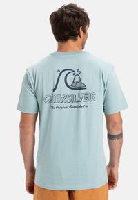 Světlemodré bavlněné tričko s grafikou na zádech, znázorňující vlnu a horu v tmavě modré barvě. Obsahuje text "Quiksilver" v tučném písmu. Krátké rukávy.