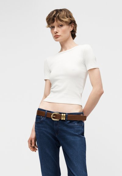 Νεαρό άτομο που φοράει στενό λευκό κοντομάνικο τοπ, μπλε τζιν και καφέ ζώνη με χρυσή αγκράφα, στέκεται μπροστά σε απλό φόντο.