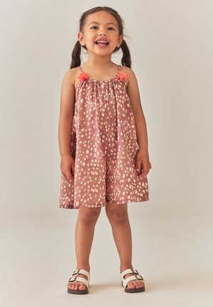 Fille souriante avec des couettes, portant une robe rose à pois avec des glands orange et des sandales blanches, debout devant un fond uni.